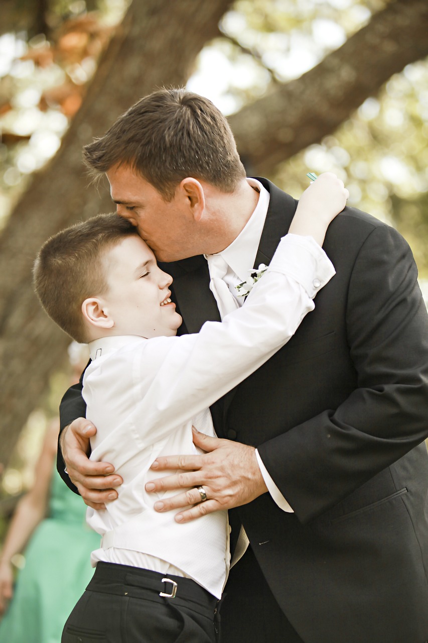 Un padre e un figlio autistico, entrambi vestiti eleganti che si abbracciano. Il padre vestito da cerimonia lo bacia sulla fronte